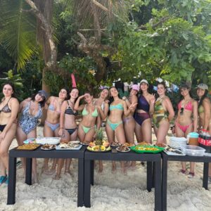 Grupo de mujeres posando en la playa detrás de una mesa con comida variada durante una reunión al aire libre